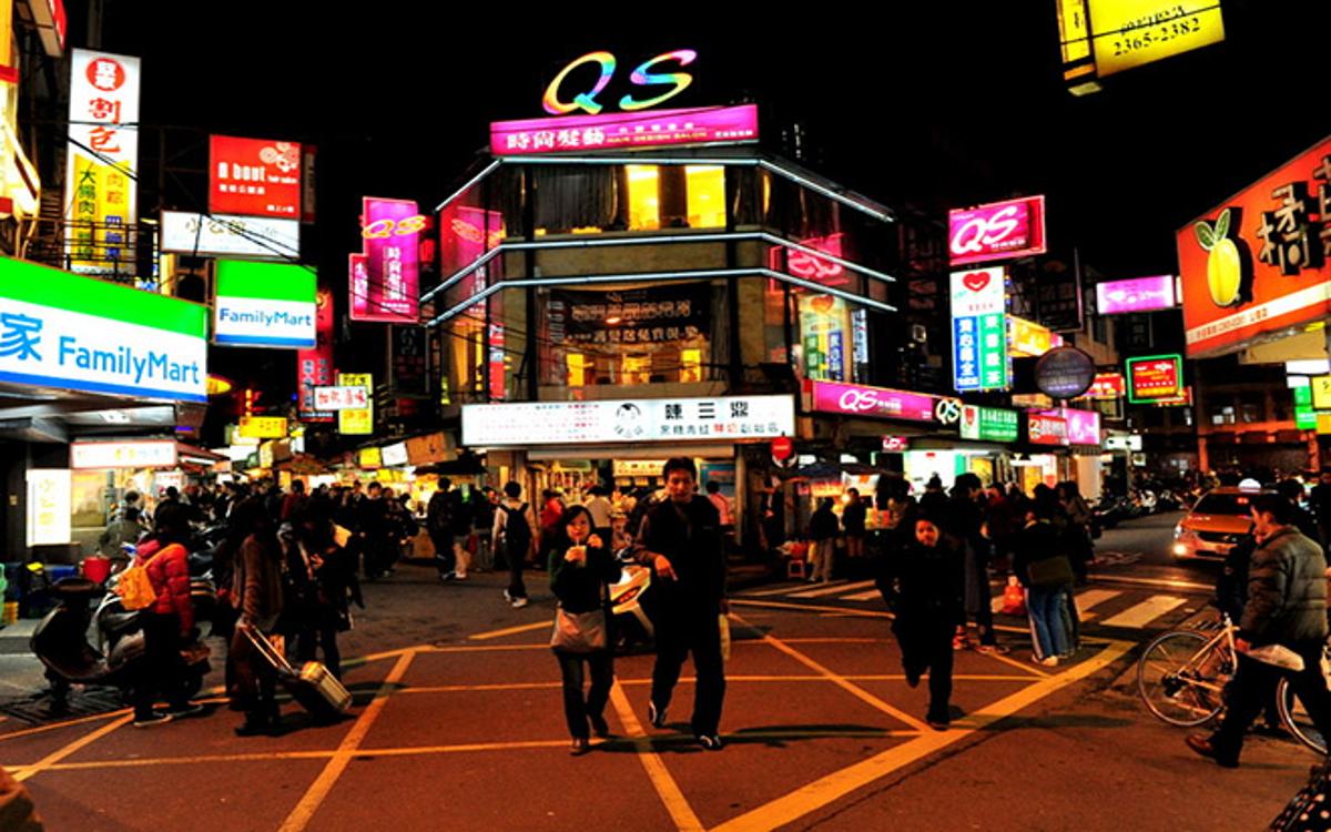 A nighttime street scene in Taipei with brightly lit neon signs and many pedestrians on the streets.