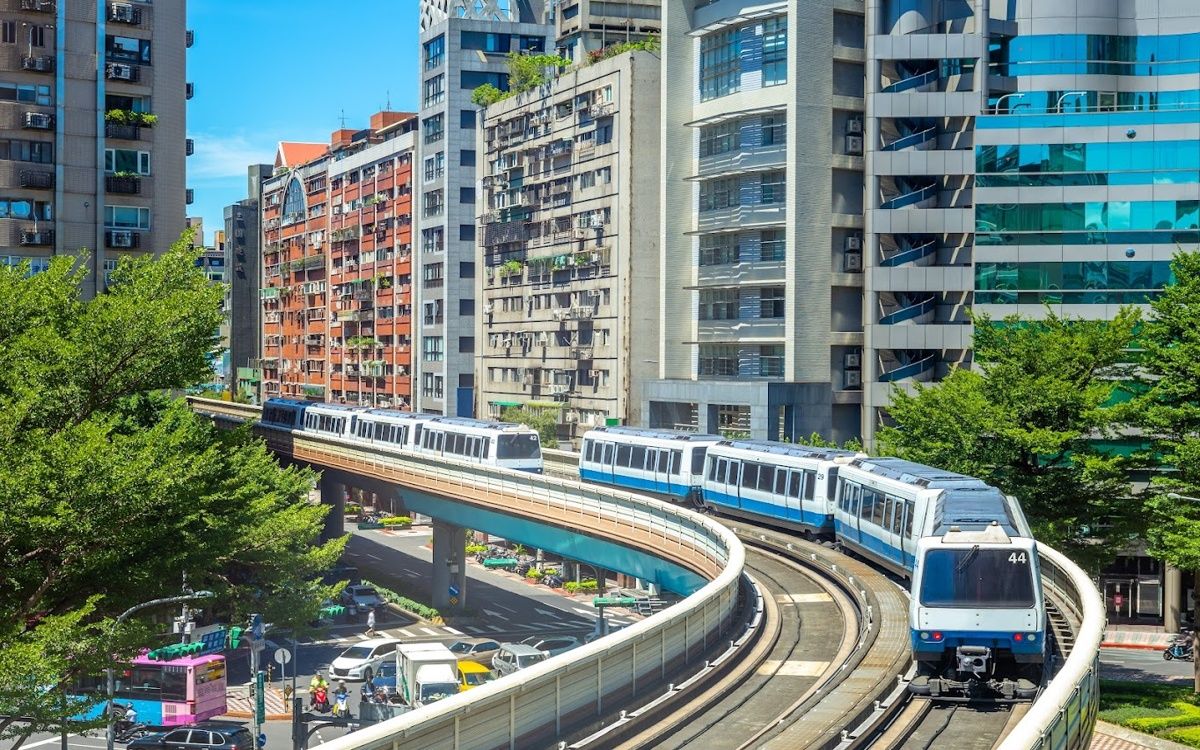 Hình ảnh tàu MRT Đài Bắc chạy trên đường ray trên cao, uốn cong giữa các tòa nhà cao tầng và cây xanh trong ngày nắng đẹp.