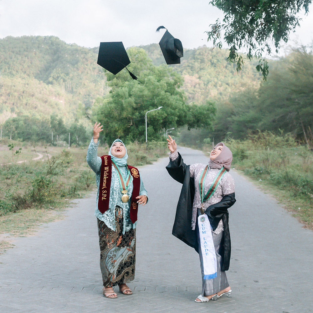 Large group of graduates throwing mortarboard caps high in air with joyful celebration under blue sky with university campus courtyard in background