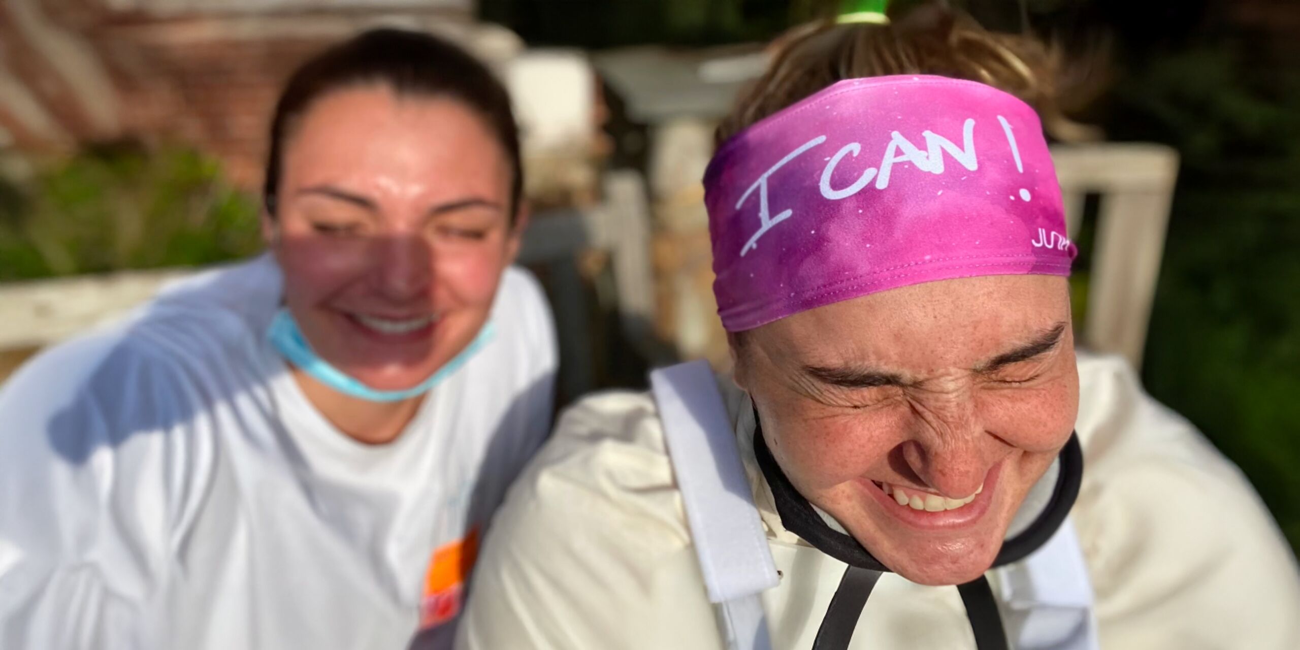 Two healthcare workers laughing outdoors, one wearing a pink headband that reads “I CAN!”
