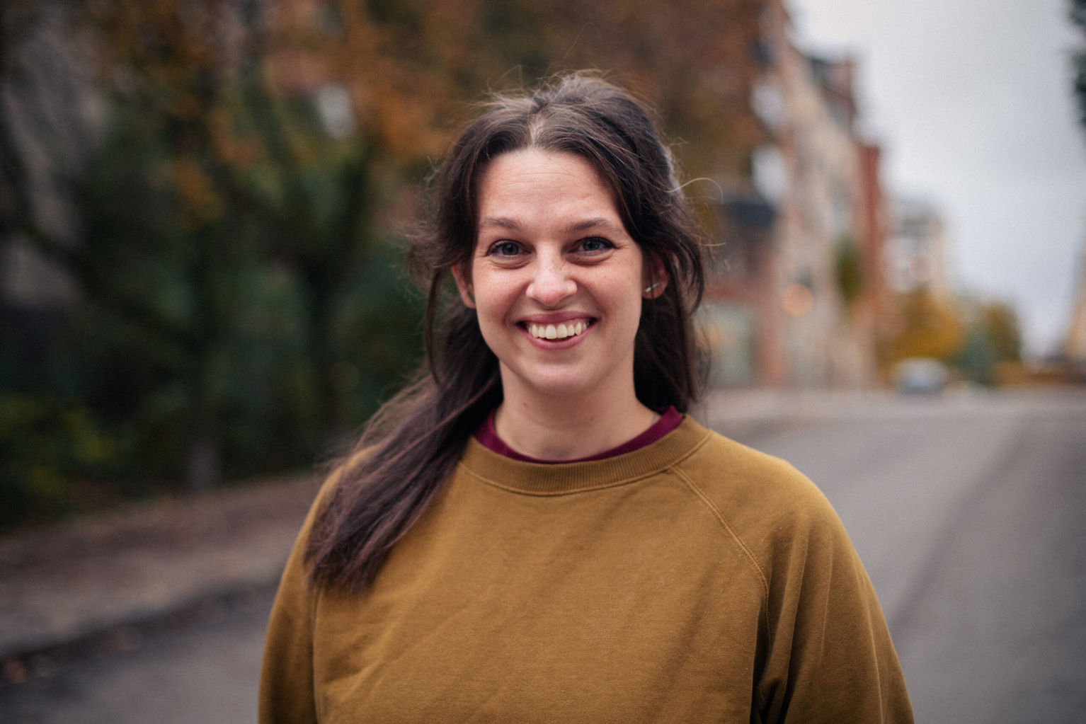 Cristine Berglund is wearing a brown sweater and smiles towards the camera. Behind her is a street lined with trees.