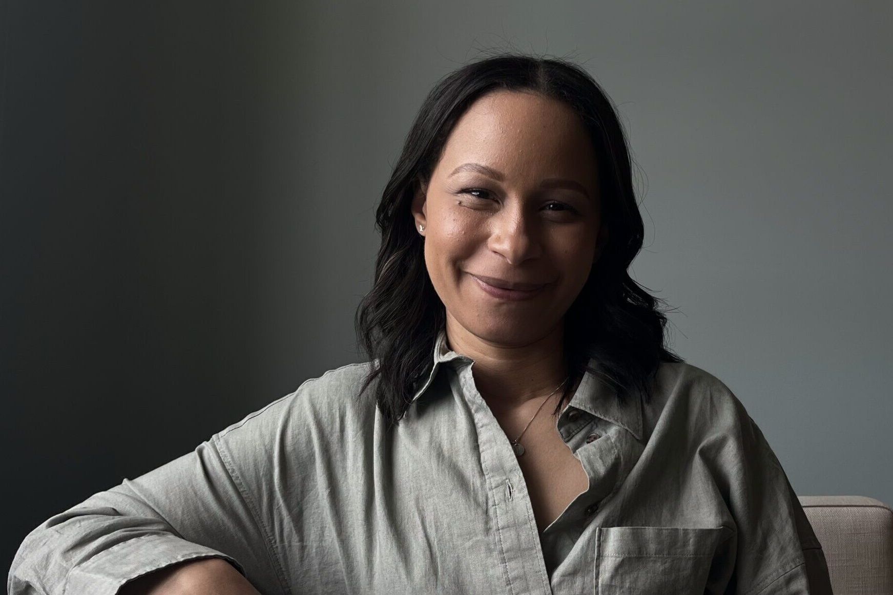 A portrait picture of Jheri Malm, smiling as she sits on the sofa inside an apartment.