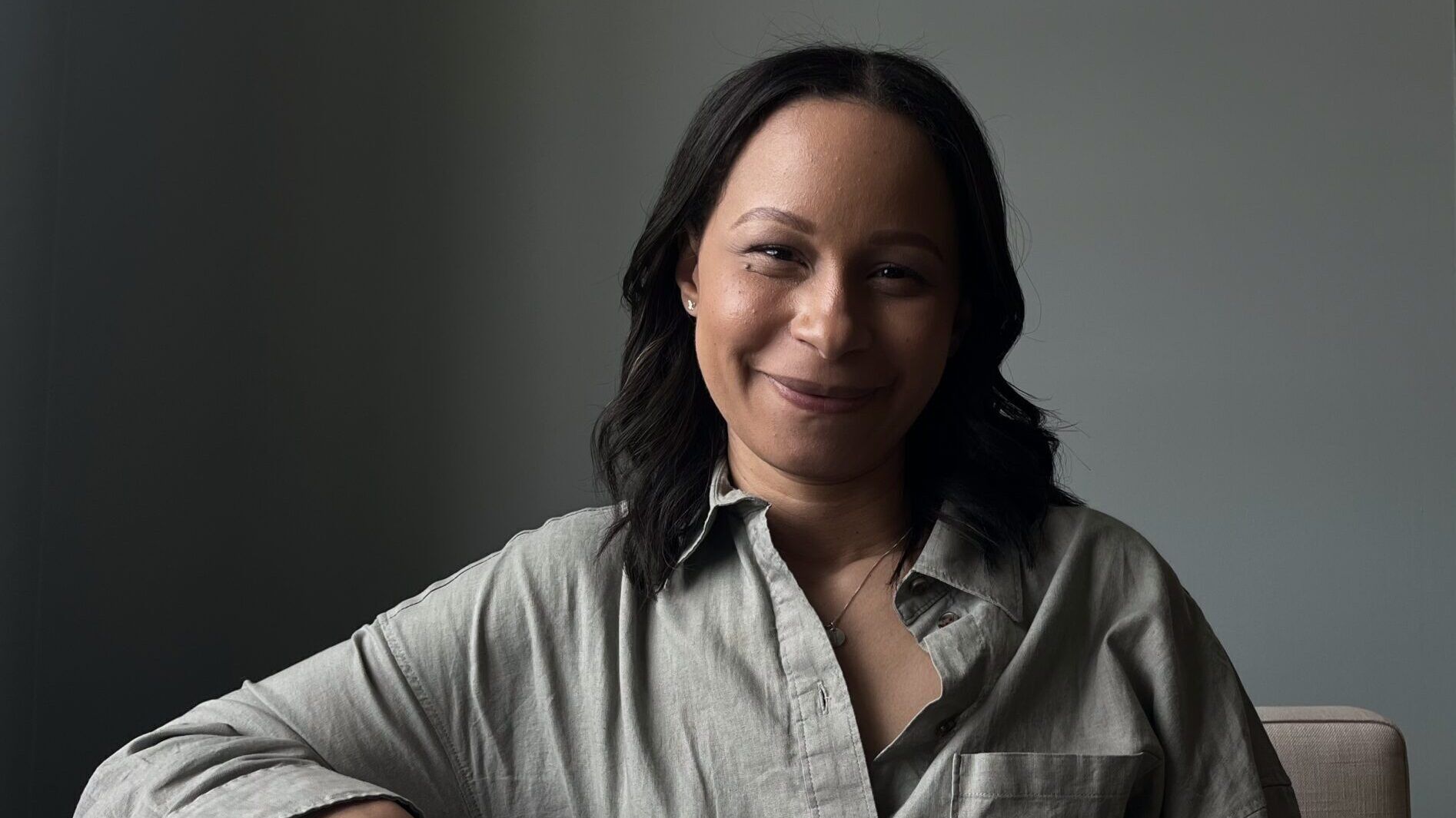 A portrait picture of Jheri Malm, smiling as she sits on the sofa inside an apartment.