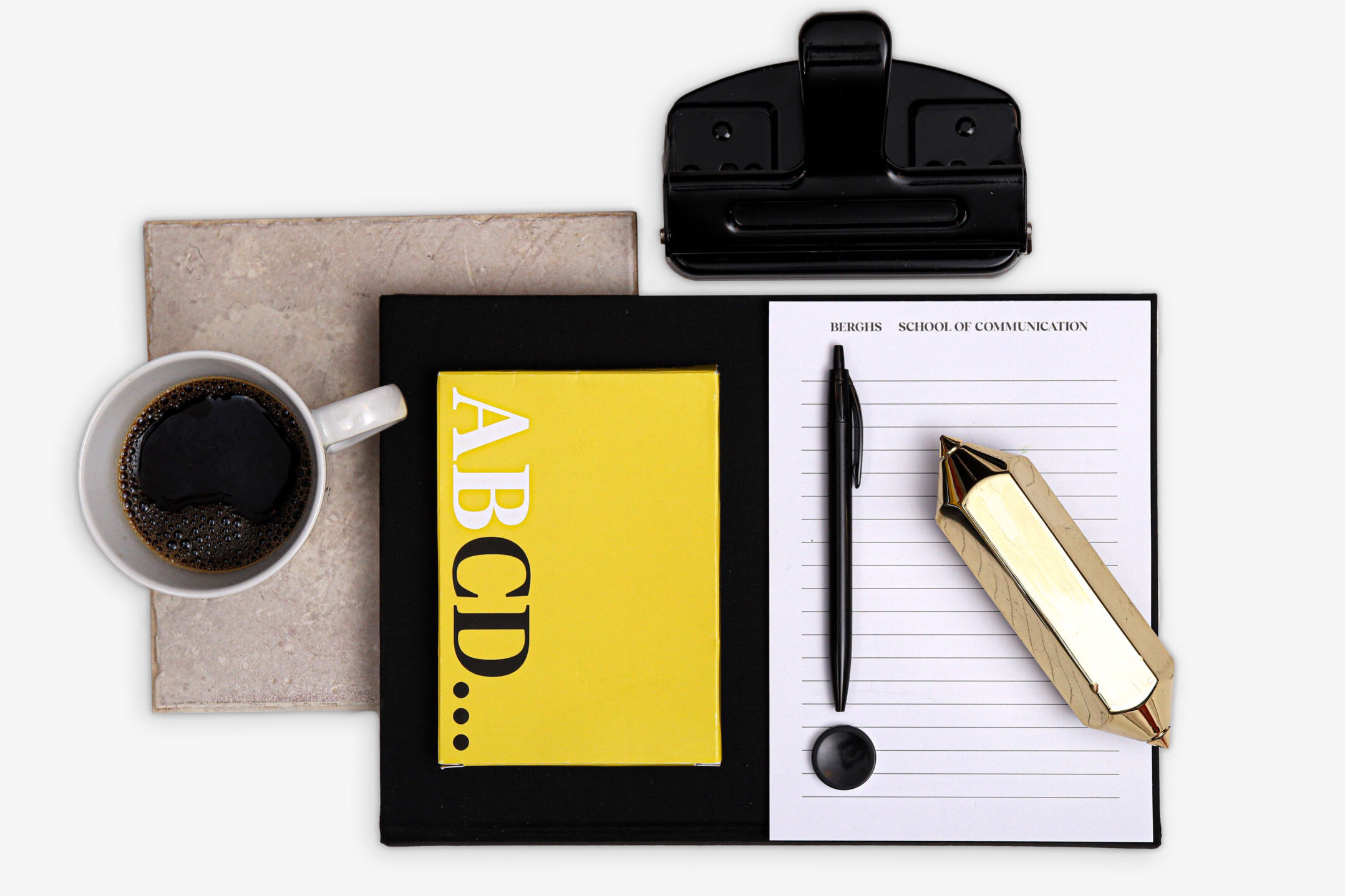 En komposition av olika föremål i gult, svart och vitt. Bilder består av en kaffekopp, block, penna och en hålslagare.