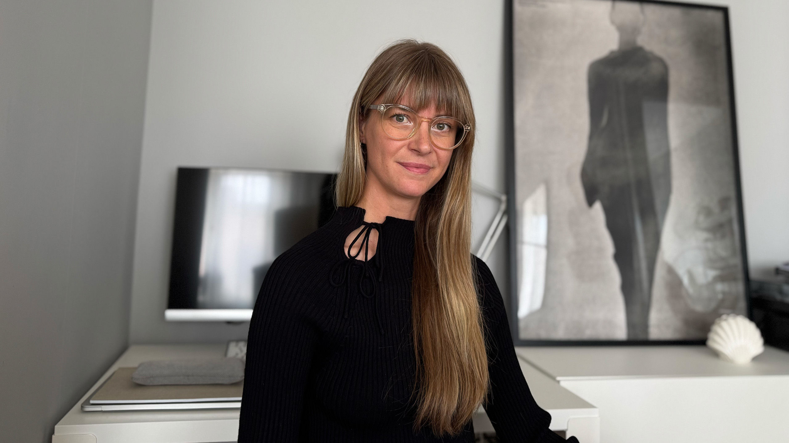Johanna Åkeson smiles towards the camera, behind her you see an Apple Desktop computer and a framed print.