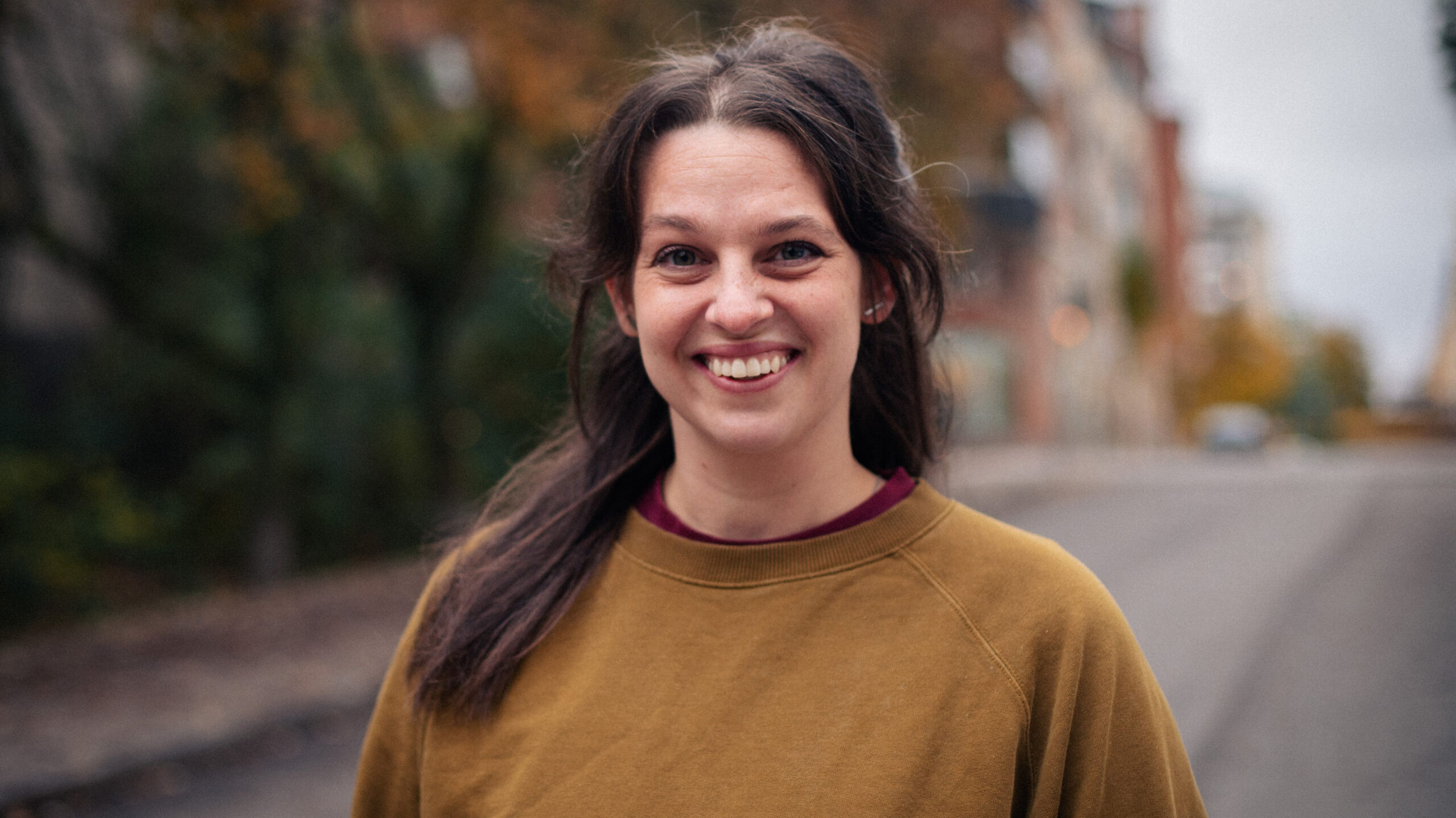 Cristine Berglund is wearing a brown sweater and smiles towards the camera. Behind her is a street lined with trees.