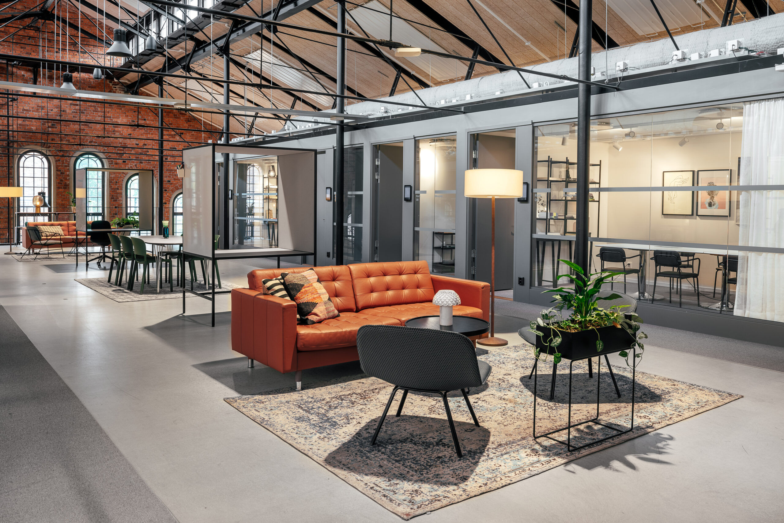 Bild på ett öppet utrymme med soffa i läder, fåtöljer samt en rad av mötesrum i bakgrunden. Industriellt tak med stålreglar samt tegelväggar.