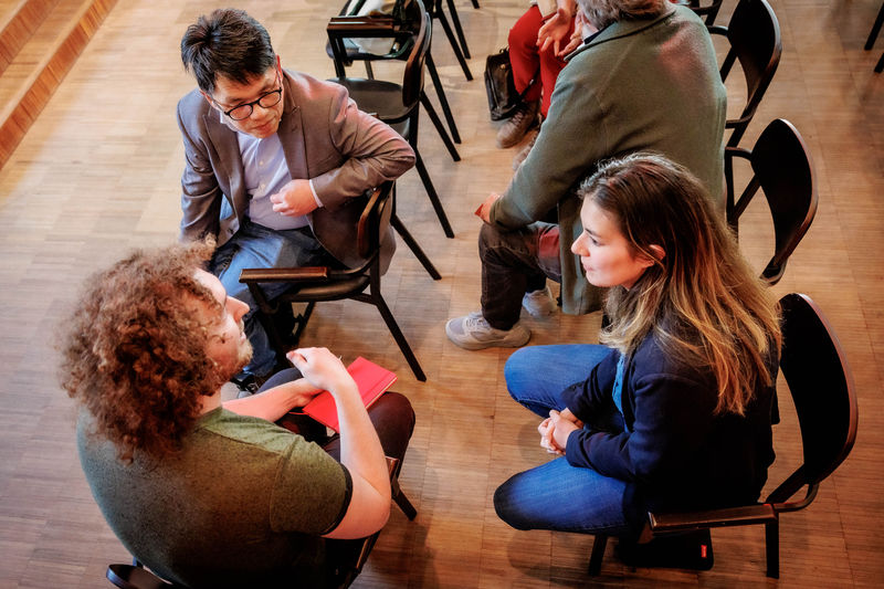 Vier mensen zitten in een kring en voeren een gesprek. Ze bevinden zich in een ruimte met houten vloer en lege stoelen op de achtergrond.