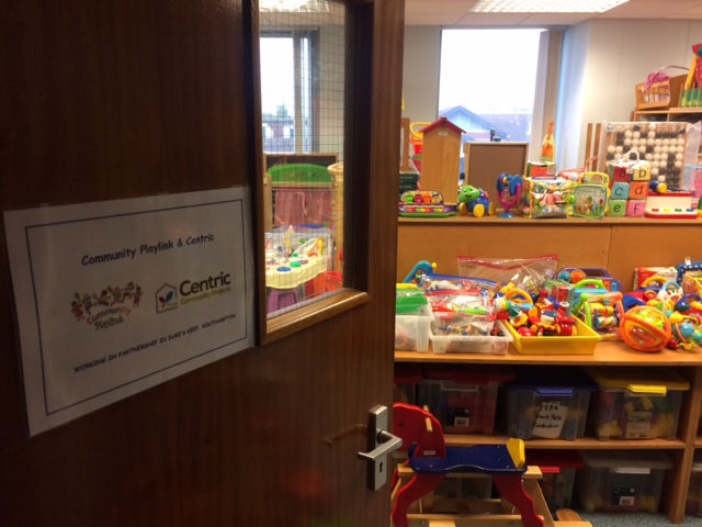 A door to the Toy Library is ajar with colourful toys on display inside. The door has a sign with the Community Playlink logo on it.