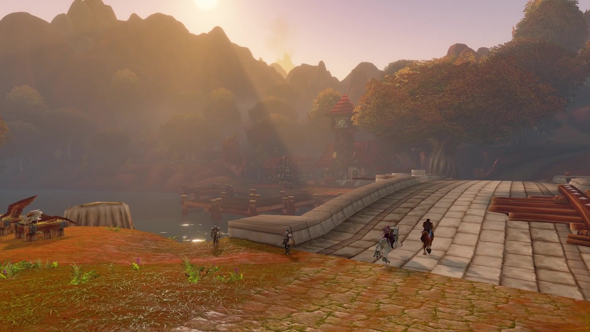 Two riders cross a stone bridge into a lakeside settlement at sunrise, wooded hills rising behind it.