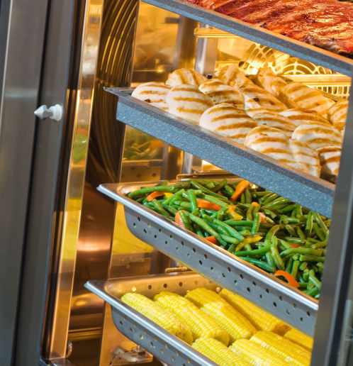 Multiple trays of food inside a commercial combi oven, including grilled chicken breasts, mixed green beans and carrots, corn on the cob, and ribs.