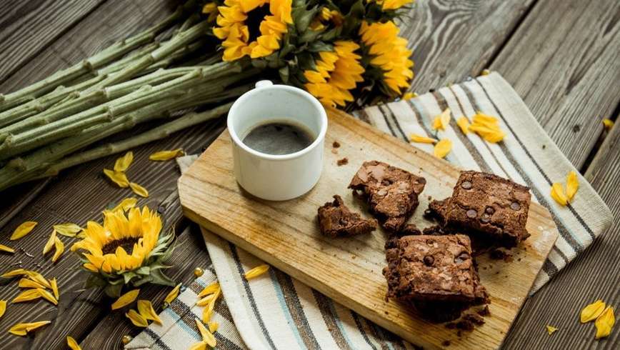 cannabis CBD brownies and cup of coffee on a cutting board. The cutting board is on a table with sunflowers beside it.