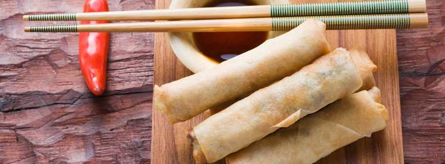 Three weed spring rolls on a wooden plate with a cup of sauce beside them. On top of the cup of sauce there is two chopsticks and a red pepper in the background
