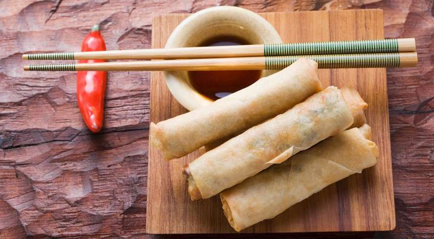 Three weed spring rolls on a wooden plate with a cup of sauce beside them. On top of the cup of sauce there is two chopsticks and a red pepper in the background