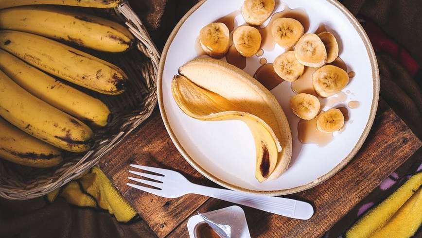 Banana on a plate that’s been cut open with another banana that still has the peel on it all of this is on a wooden cutting board with other bananas in the background