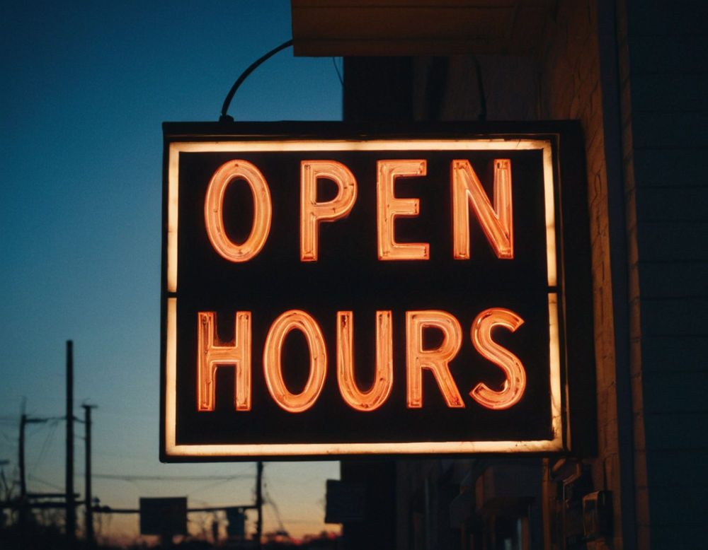 A sign displaying 'Open 24 Hours' illuminated by the warm glow of a streetlamp outside a laundromat during dusk.