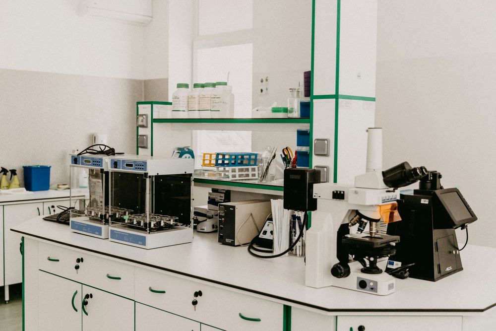 A peptide laboratory workspace featuring a white countertop with various scientific equipment, including two analytical devices, a microscope, and electronic instruments, along with shelves stocked with lab supplies and tools, all set against a light-colored wall.