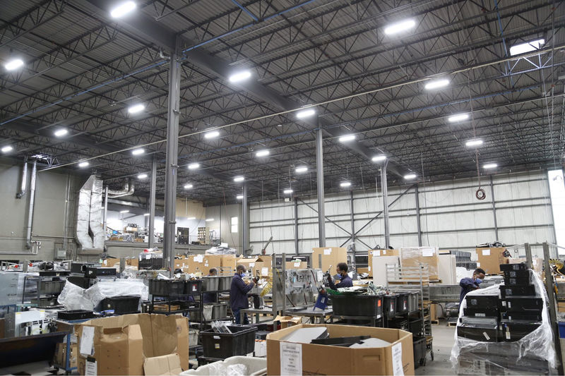 A spacious industrial warehouse with high ceilings, featuring bright LED lighting. Workers are seen handling equipment and materials among various storage bins and cardboard boxes, creating a busy and organized environment.