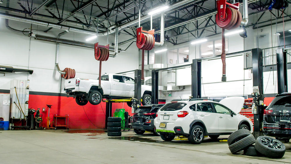 A modern auto repair shop featuring several vehicles, including a lifted white truck and a variety of parked cars. The interior has red and white walls, hanging tools, and equipment, with tires on the floor and a mechanic visible in the background.