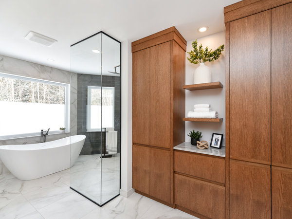 Luxurious modern bathroom pairing warm wood full-height cabinetry with white marble tile floors and walls, featuring a freestanding soaking tub and glass shower enclosure.