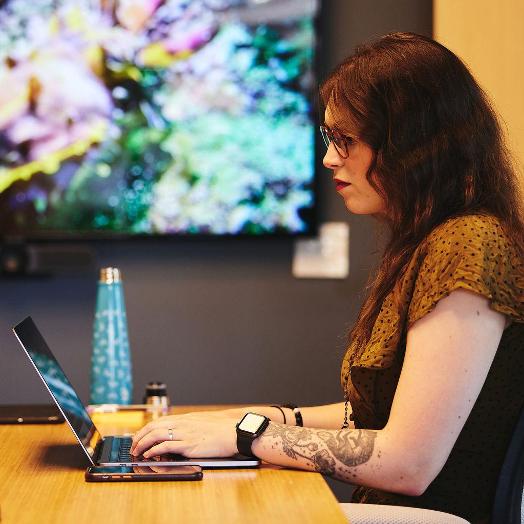 Emily Morehouse, Co-founder and Director of Engineering at Cuttlesoft, works intently on Ruby development at her workspace, embodying the company's artistic approach to software engineering. The thoughtfully composed image shows her focused on a MacBook Pro, with a vibrant digital display in the background creating an artistic atmosphere that reflects her unique background bridging creative arts and technical expertise. Wearing a patterned yellow blouse and modern tech accessories including an Apple Watch, she represents the contemporary face of Ruby and Rails development. The professional yet creative setting showcases Cuttlesoft's approach to custom software development, where artistic sensibility meets technical precision. Emily's workspace, featuring a sleek laptop, ergonomic setup, and personal touches like a designer water bottle, illustrates the company's investment in both cutting-edge tools and developer comfort. This environment captures Cuttlesoft's philosophy of crafting elegant Ruby solutions, combining technical excellence with creative problem-solving for enterprise clients. The image emphasizes the human element in software development while highlighting the company's capabilities in Ruby, Rails, and full-stack web development services.