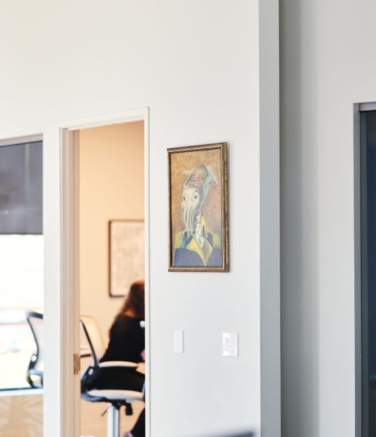 A glimpse into Cuttlesoft's workspace shows their company mascot - an artistic interpretation of a cuttlefish - displayed prominently on the wall, representing their philosophy of adaptability in software development. Through the doorway, team members are visible collaborating in a modern office setting equipped for full-stack development work. The clean, minimalist office design reflects Cuttlesoft's approach to creating elegant solutions for enterprise clients across healthcare, government, and technology sectors. The cuttlefish artwork, central to the company's identity, symbolizes their ability to adapt and integrate with client teams while delivering custom software solutions in Python, Ruby, React, and mobile development. This environment embodies their core value of 'Put Knowledge Into Practice' while maintaining their commitment to both technical excellence and human-centered development.