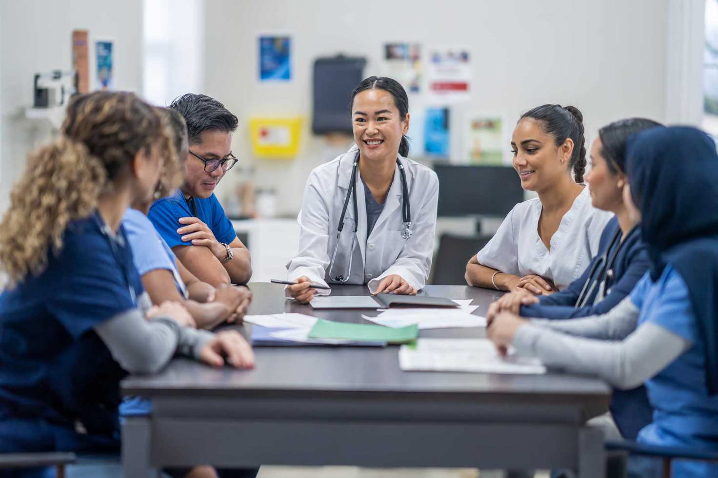 This image showcases a professional and engaging scenario typical at healthcare educational workshops, aligning perfectly with the platform developed by Cuttlesoft for Zygo Health. It features a diverse group of medical professionals gathered around a table in a bright, modern classroom setting. The central figure, a confident female doctor wearing a white coat, leads the discussion, drawing attention with her engaging demeanor. She is surrounded by colleagues in various healthcare uniforms, indicating a collaborative and interdisciplinary learning environment.