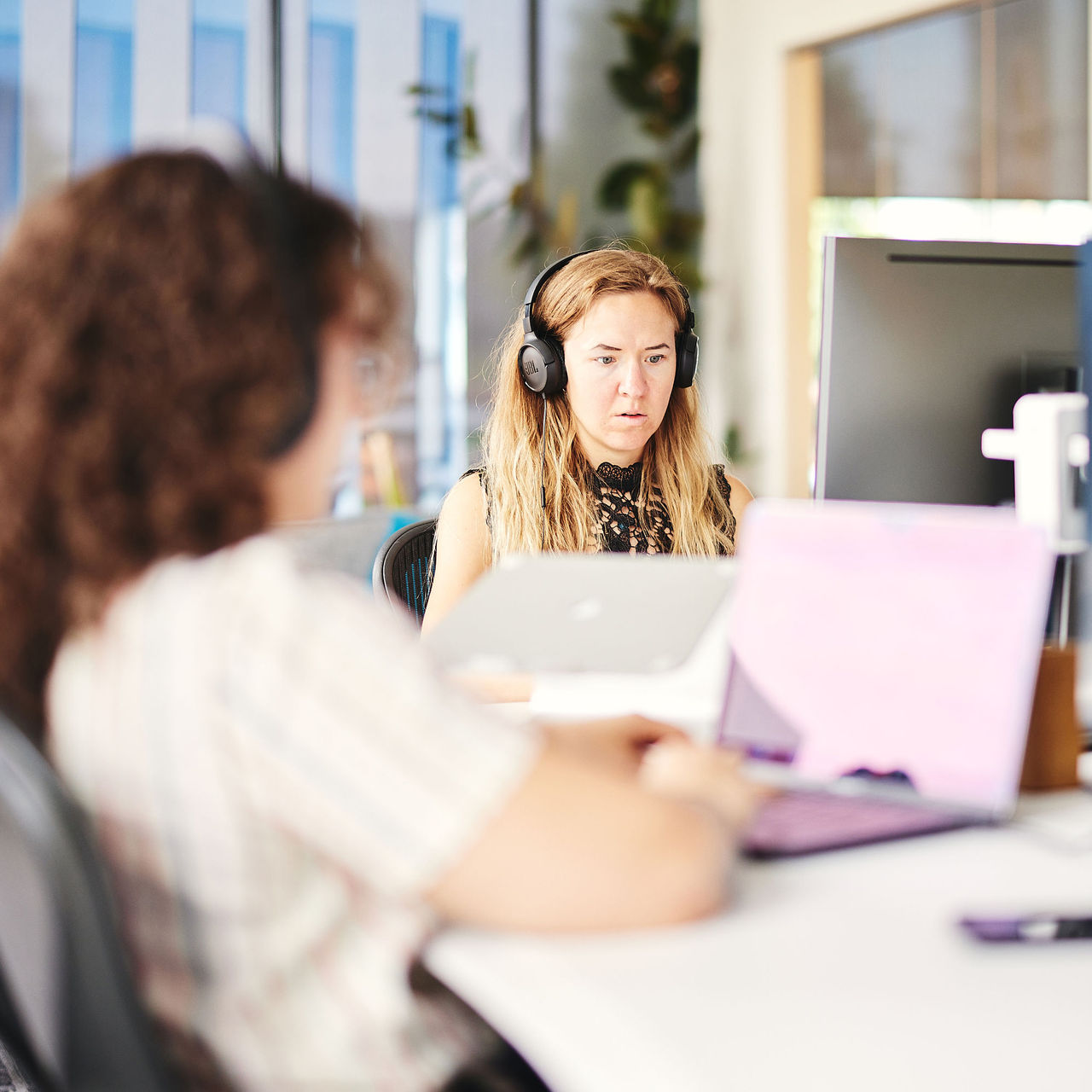 A software engineer wearing headphones is focused intently on her development work at Cuttlesoft's modern office space. Sara, a Node.js expert, sits at a collaborative workspace equipped with dual monitors and a laptop, demonstrating the company's hands-on approach to custom software development. She wears a black lace-detailed sleeveless top and over-ear headphones, suggesting both professionalism and deep concentration while coding. The bright, open-plan office features natural lighting through large windows and decorative plants, reflecting Cuttlesoft's commitment to creating an optimal environment for technical innovation. In the foreground, a team member is visible, indicating the collaborative nature of their Node.js development services. The scene captures the essence of modern software development - combining individual focus time with team collaboration. The setup includes industry-standard development tools and ergonomic furniture, highlighting Cuttlesoft's investment in both their team's productivity and comfort. This working environment represents the company's approach to enterprise-grade Node.js development, backend architecture, and full-stack JavaScript solutions, where dedicated engineers like Sara craft scalable, efficient code for custom business applications.