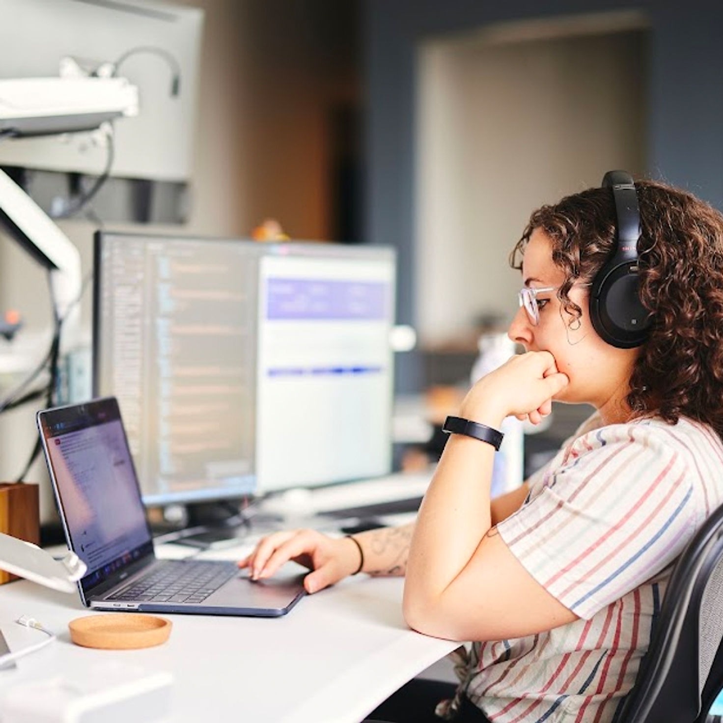 Marisa, a focused software developer engaged in custom web application development at Cuttlesoft. This professional is captured in a well-equipped workspace featuring multiple screens displaying sophisticated code, one showcasing Angular, another focused on MongoDB, and an IDE with Node.js scripts evident. The developer, wearing a comfortable striped T-shirt and headphones, mirrors the concentration and expertise required to handle complex educational software solutions and data analytics platforms. This image clearly portrays the intense focus and technical skill involved in developing user-friendly, efficient educational technology solutions in the EdTech sector, enhancing the ability of institutions to harness big data for impactful insights.