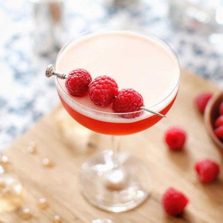 Três taças contendo o drink Clover Club, com framboesas espetadas em palitos apoiados nas bordas, rodeadas por mais framboesas, uma garrafa de gin Beefeater e outros objetos.