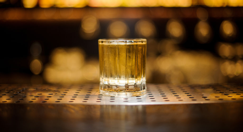 Imagem de um copo de vidro com o drink Rusty Nail sobre a bancada de um bar, com objetos desfocados ao fundo.