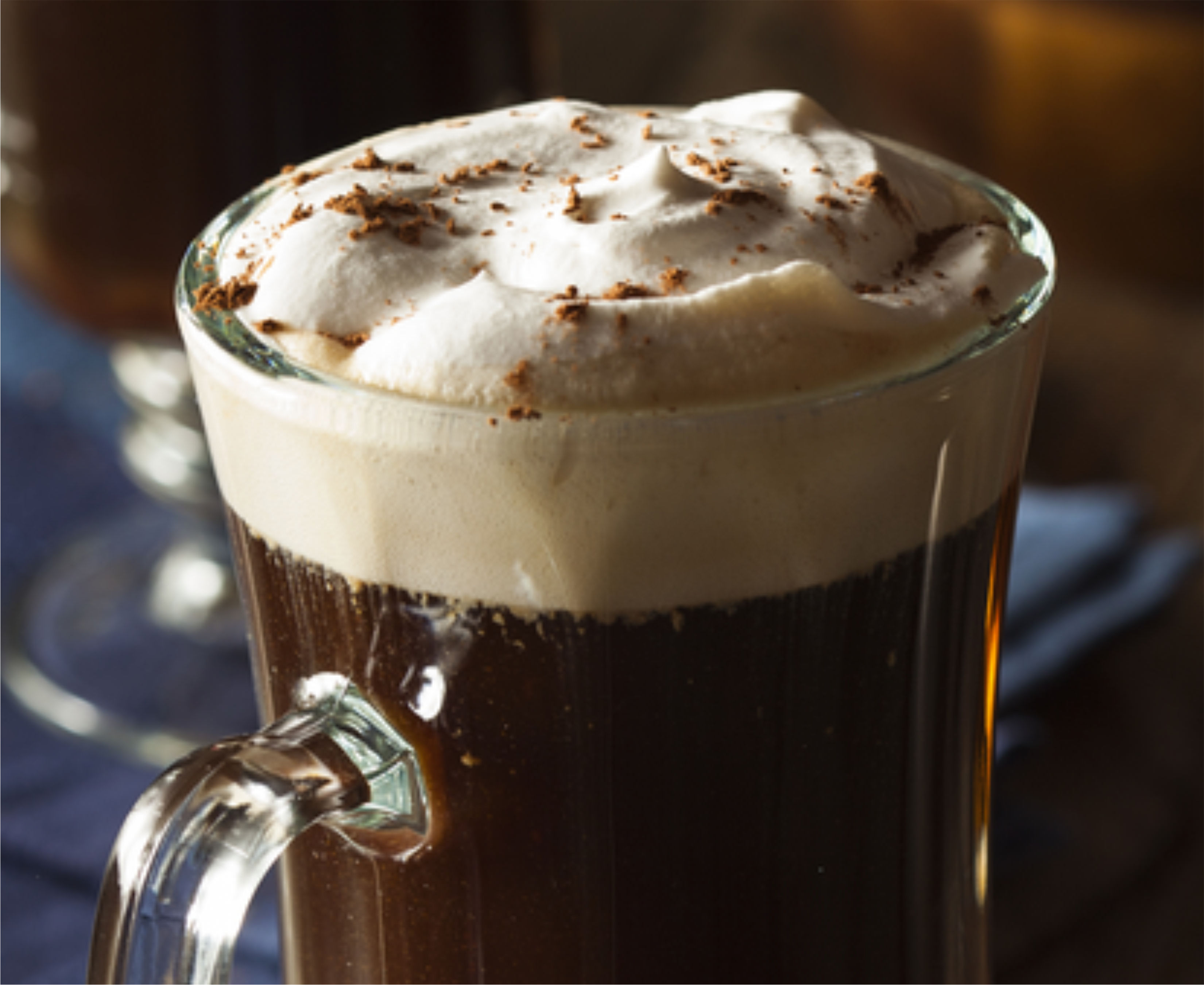 Imagem de uma caneca de vidro com o drink de café com whisky e chantilly, sobre um guardanapo de pano azul, ao lado de outras louças, potes e colheres.
