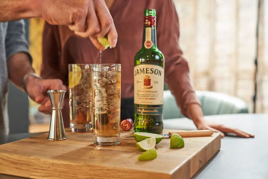 Cocktail preparation featuring a person squeezing fresh lime juice into a glass. A bottle of Jameson whiskey, a jigger, and lime wedges are placed on a wooden cutting board.