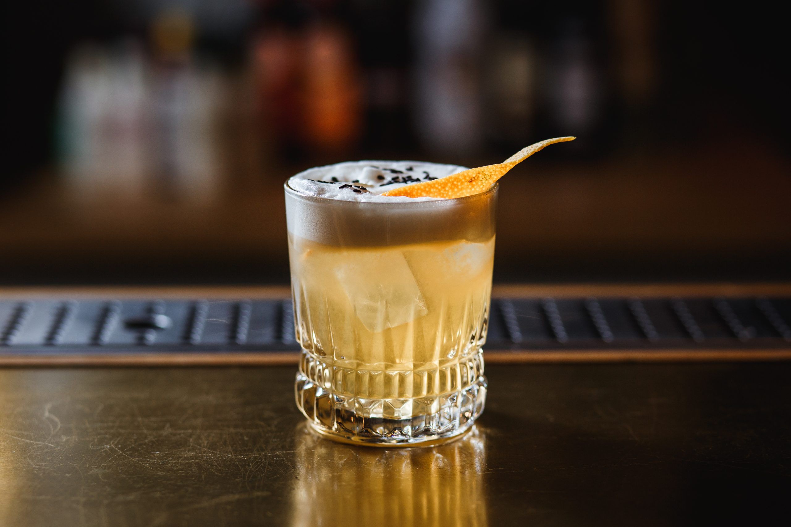 a light orange cocktail in an old fashioned glass, covered with egg white foam. decorated with sesame seeds and orange peel. ice cubes inside. selective focus.