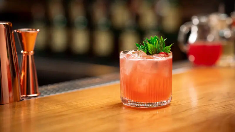 A vibrant pink cocktail served over ice in a textured glass, garnished with fresh mint and a strawberry, set on a polished wooden bar with copper bar tools and a blurred background.