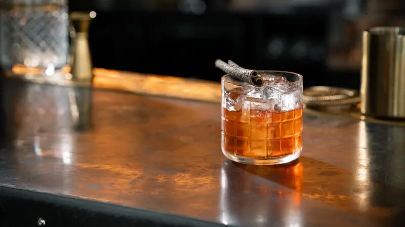 A glass of whiskey with ice, garnished with a cinnamon stick, sits on a dimly lit bar counter with warm reflections.