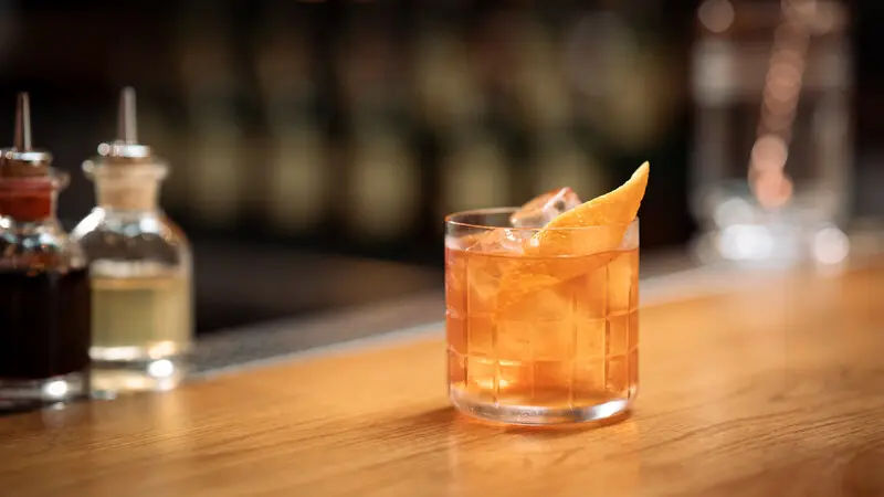 An Old Fashioned cocktail with ice and an orange peel garnish sits on a wooden bar counter.
