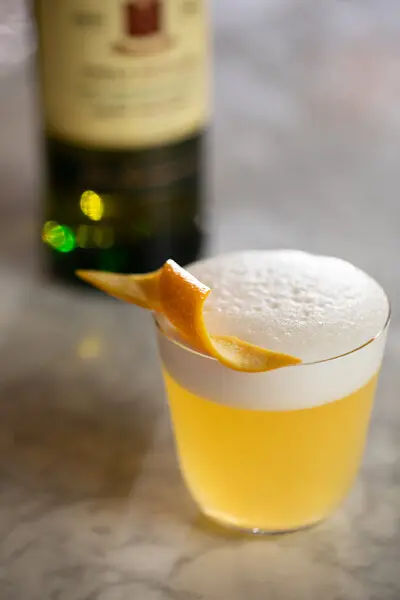 A whiskey sour cocktail with a frothy top, garnished with an orange peel, sits on a marble surface with a blurred bottle of Jameson in the background.