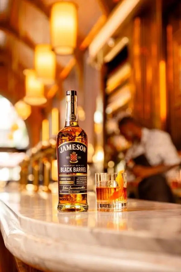 a bottle of jameson black barrel whiskey and a cocktail garnished with an orange twist on a bar counter, with a bartender blurred in the background