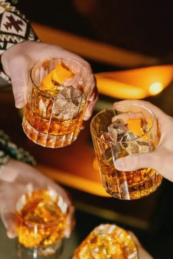 two hands clinking glasses of whiskey with ice and orange peels, with additional drinks reflected on a glass surface under warm lighting