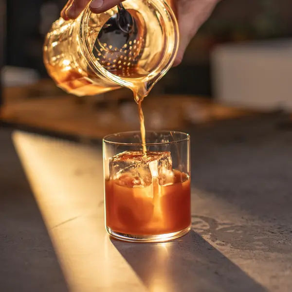 A hand pours a chocolate old fashioned cocktail from a mixing glass into a short tumbler with a large ice cube, illuminated by warm sunlight on a smooth countertop.
