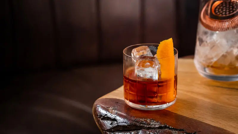 A rich amber cocktail with ice and an orange peel garnish is served in a short glass on a rustic wooden table, with a dark leather background and a mixing glass nearby.