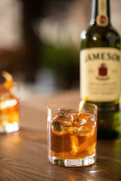 An Old Fashioned cocktail with ice and an orange twist sits on a wooden table with a blurred Jameson Irish Whiskey bottle in the background