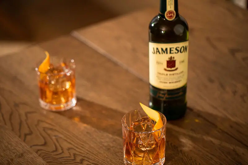 A bottle of Jameson Irish Whiskey sits on a wooden table next to two old fashioned cocktails garnished with orange peels, served in short, faceted glasses over ice.