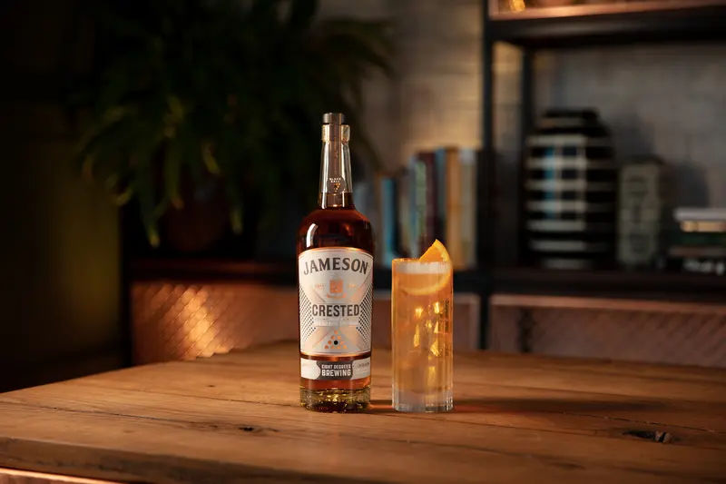 A bottle of Jameson Crested Whiskey stands beside a tall glass of cocktail garnished with an orange wedge, set on a wooden table in a warmly lit room with bookshelves and decorative items in the background.
