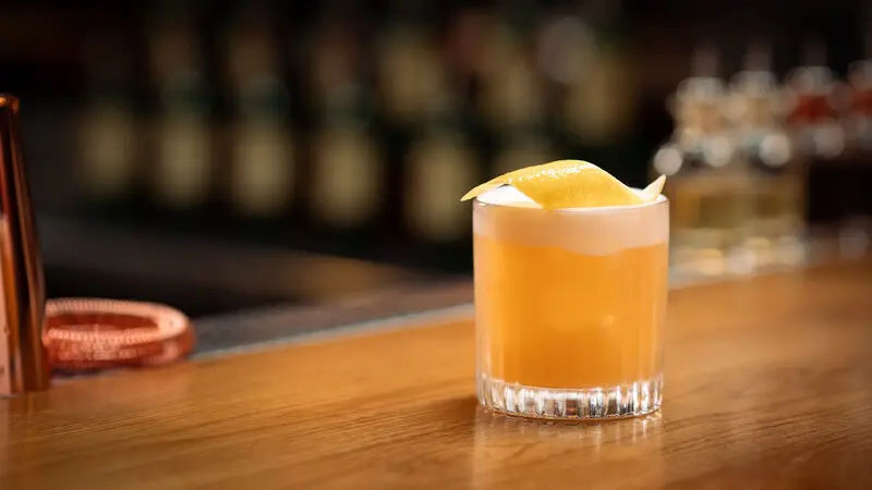 A whiskey sour cocktail with a frothy top and a lemon peel garnish is served in a short glass on a polished wooden bar, with a blurred background of bottles and bar tools.
