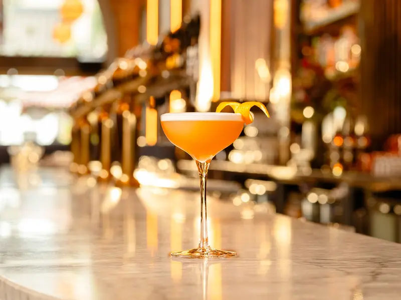 A whiskey sour cocktail in a coupe glass garnished with a citrus twist, set on a polished bar in a warmly lit venue.