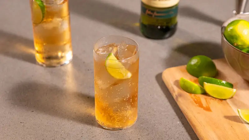 Tall glass of Jameson, ginger ale, and lime served over ice with a lime wedge garnish, placed on a grey countertop alongside a cutting board with sliced limes and a metal bowl of whole limes.