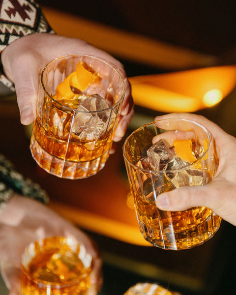 Two hands clinking Old Fashioned cocktails with orange peel garnish over ice