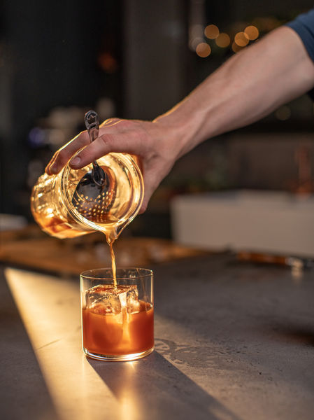 Hand pouring a rich, dark cocktail from a mixing glass intoa. rocks glass over ice in a warm, softly lit bar setting.